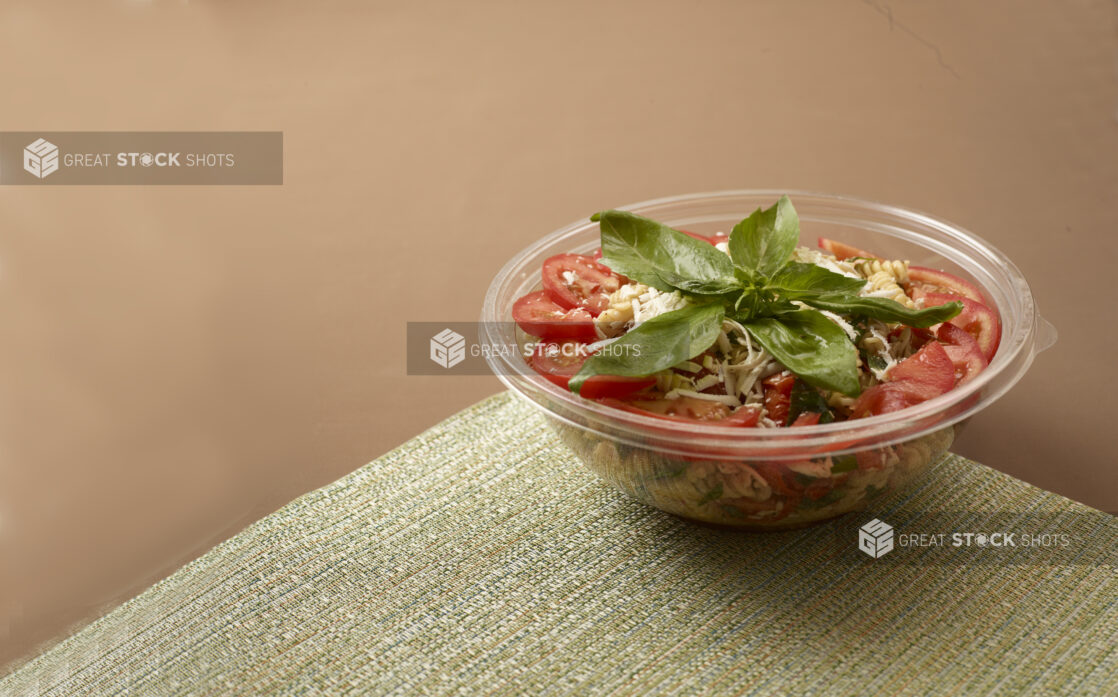 clear plastic take out bowl of a pasta salad topped with basil and sliced tomato