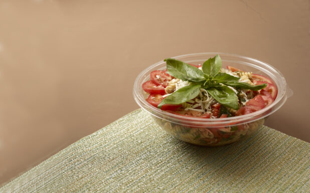clear plastic take out bowl of a pasta salad topped with basil and sliced tomato