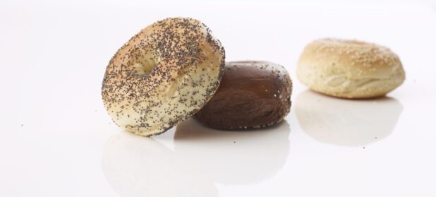 poppy seed bagel resting on a pumpernickel bagel with a sesame seed bagel out of focus in the back ground