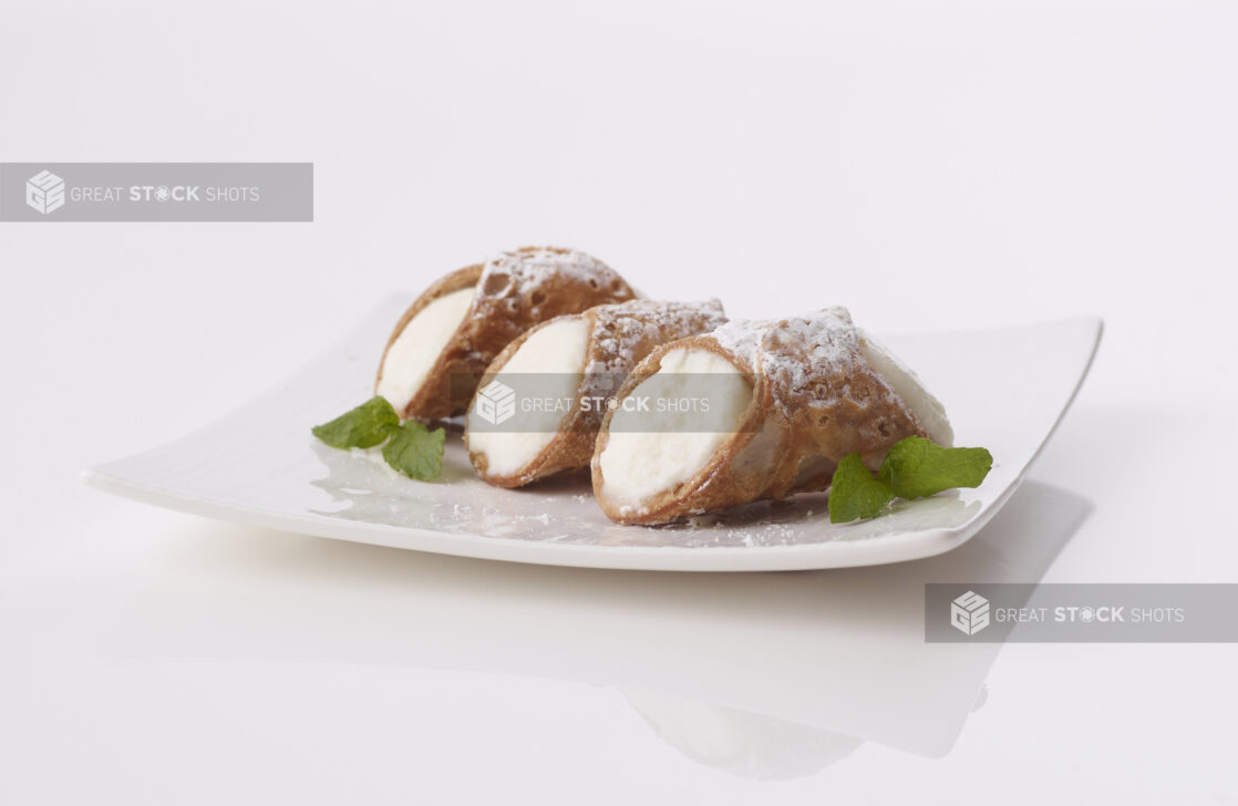 3 italian cannolis filled with ricotta cheese on a white plate sprinkled with powdered sugar garnished with mint leaves