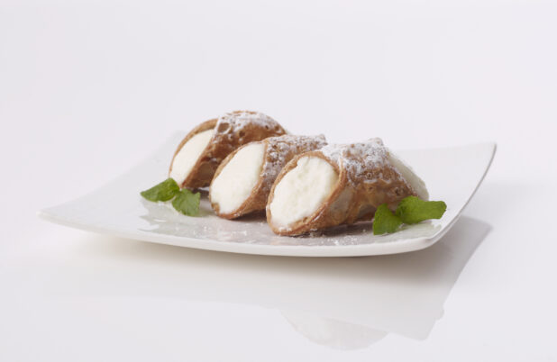 3 italian cannolis filled with ricotta cheese on a white plate sprinkled with powdered sugar garnished with mint leaves