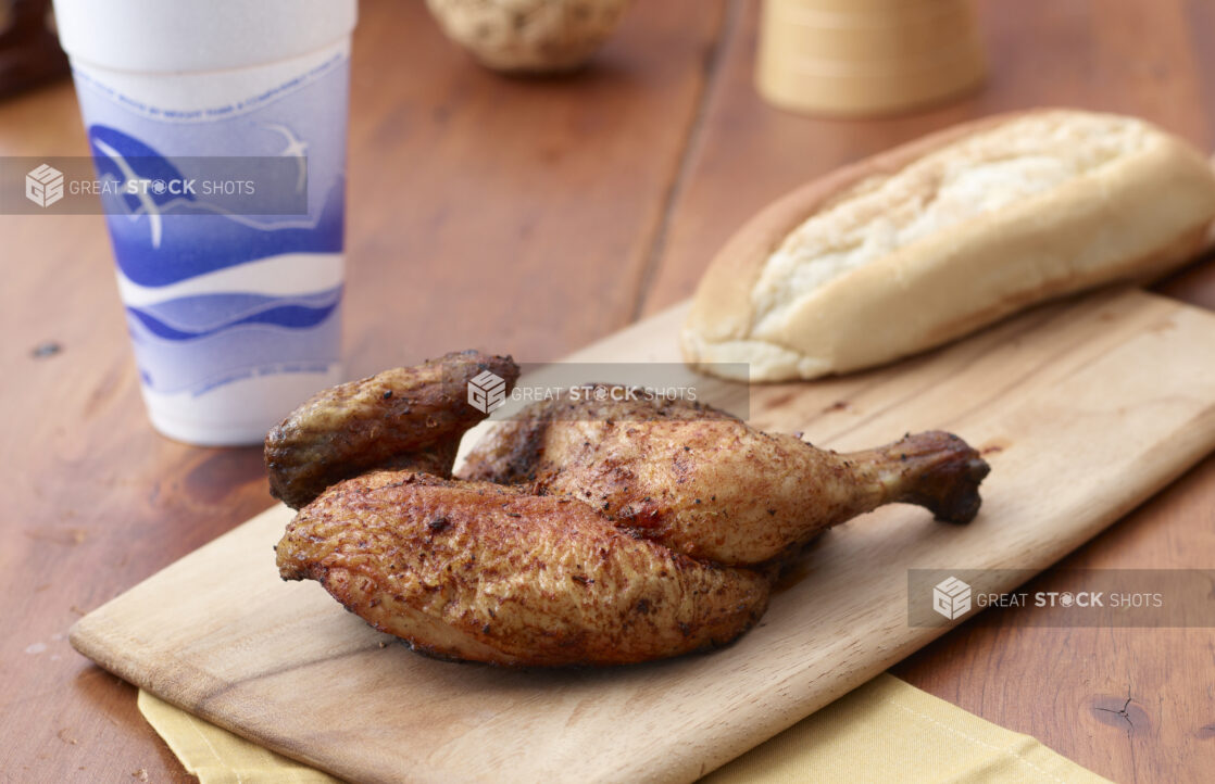 Rotisserie Chicken Leg on a Wooden Cutting Board with a Fountain Soda and Dinner Roll on a Wooden Table in an Indoor Setting