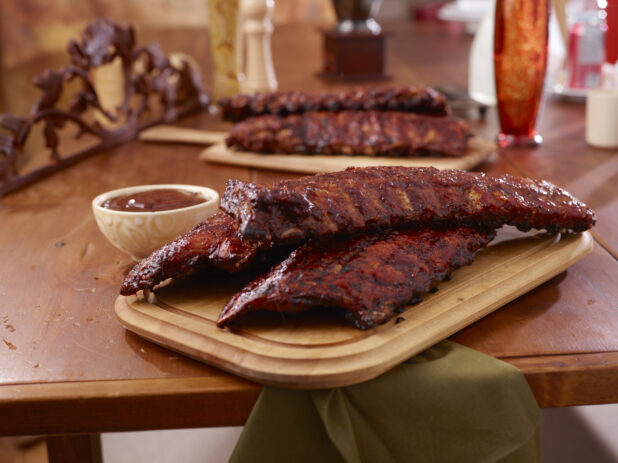 A Wooden Platter of Whole Racks of Ribs and a Side of Sauce on a Wooden Table in an Indoor Setting - Variation