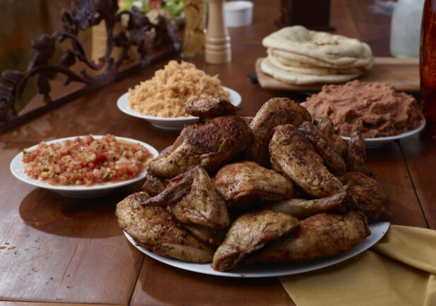A Platter of Rotisserie Chicken Legs and Side Dishes of Spanish Rice, Tomato Salsa, Refried Beans and Baguettes on a Wooden Table in an Indoor Setting