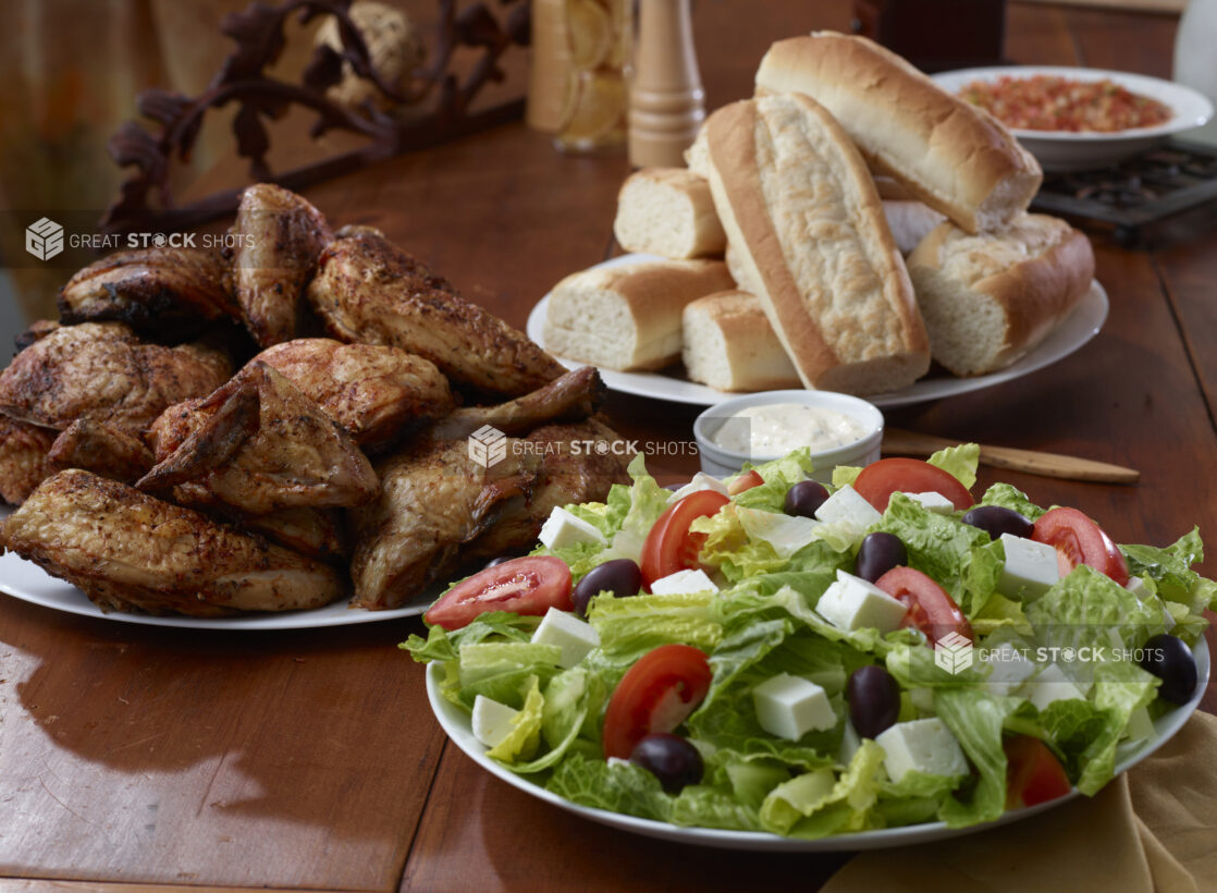 A Family Meal Consisting of Greek Salad, Rotisserie Chicken and White Bread Rolls on a Wooden Table in a Restaurant Setting