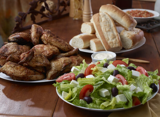 A Family Meal Consisting of Greek Salad, Rotisserie Chicken and White Bread Rolls on a Wooden Table in a Restaurant Setting