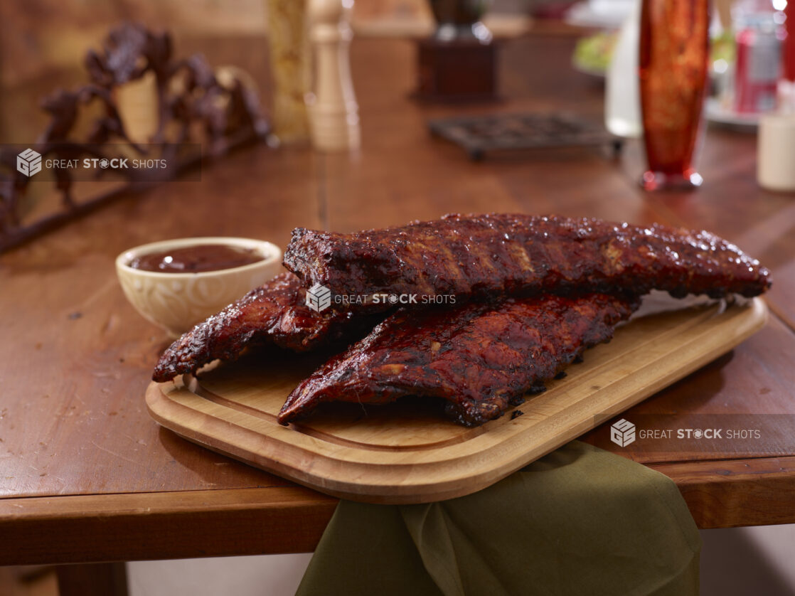 A Wooden Platter of Whole Racks of Ribs and a Side of Sauce on a Wooden Table in an Indoor Setting