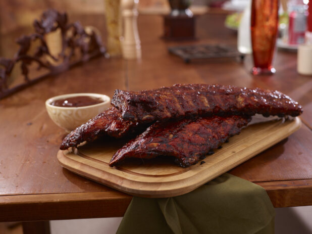 A Wooden Platter of Whole Racks of Ribs and a Side of Sauce on a Wooden Table in an Indoor Setting