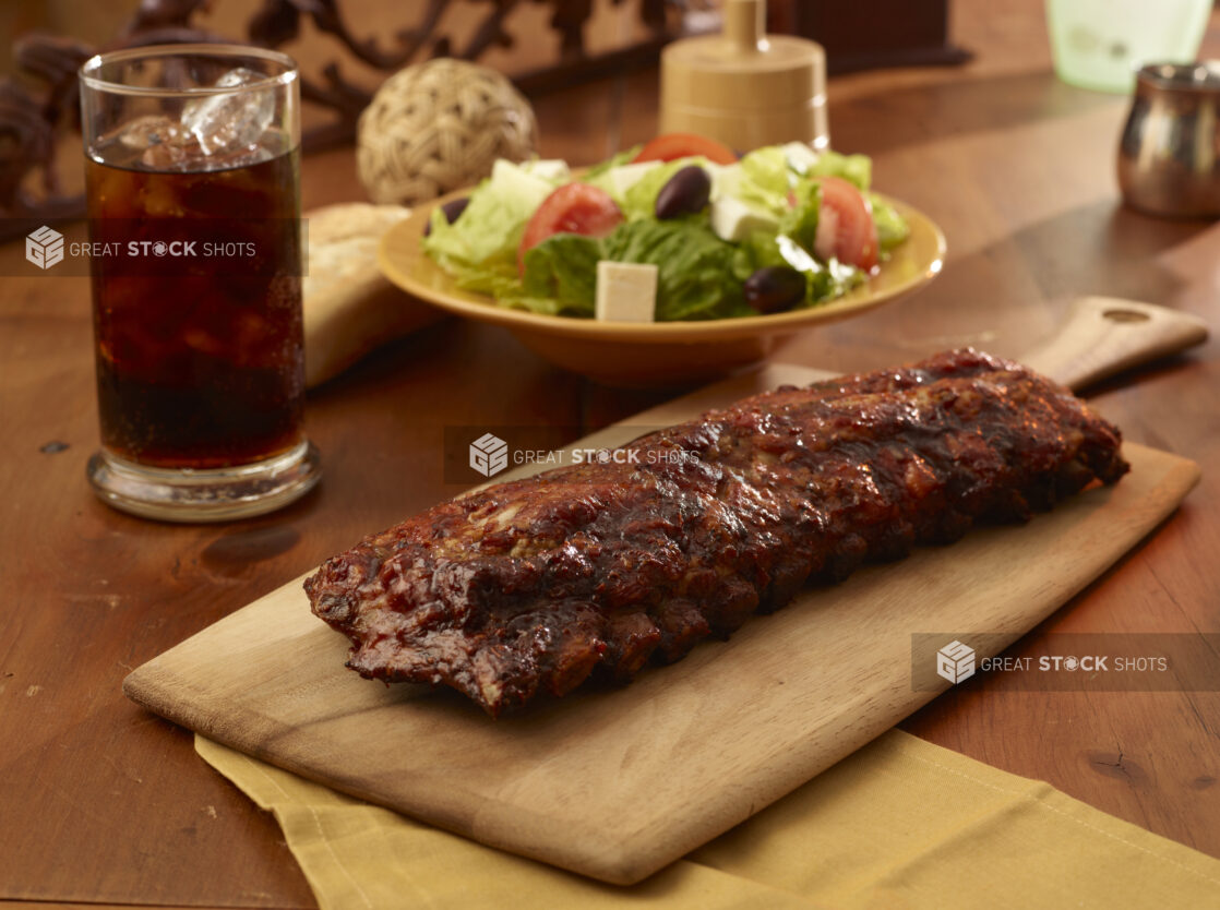 Whole Rack of Barbecue Ribs on a Wooden Cutting Board with a Glass of Soda and a Bowl of Fresh Greek Salad on a Wooden Table in an Indoor Setting