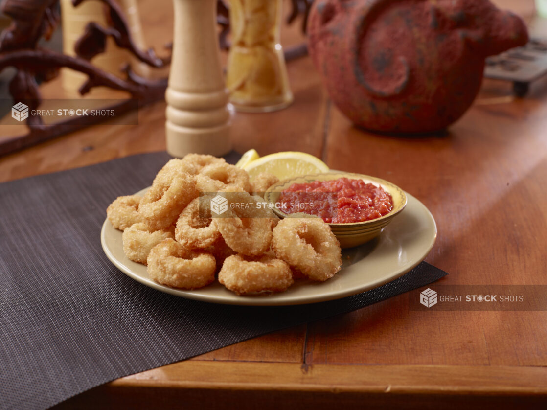 A Plate of Deep Fried Breaded Calamari Served with Cocktail Sauce and Lemon Wedges on a Wooden Table Surface in an Indoor Setting