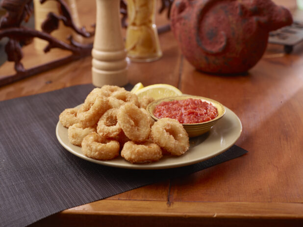 A Plate of Deep Fried Breaded Calamari Served with Cocktail Sauce and Lemon Wedges on a Wooden Table Surface in an Indoor Setting