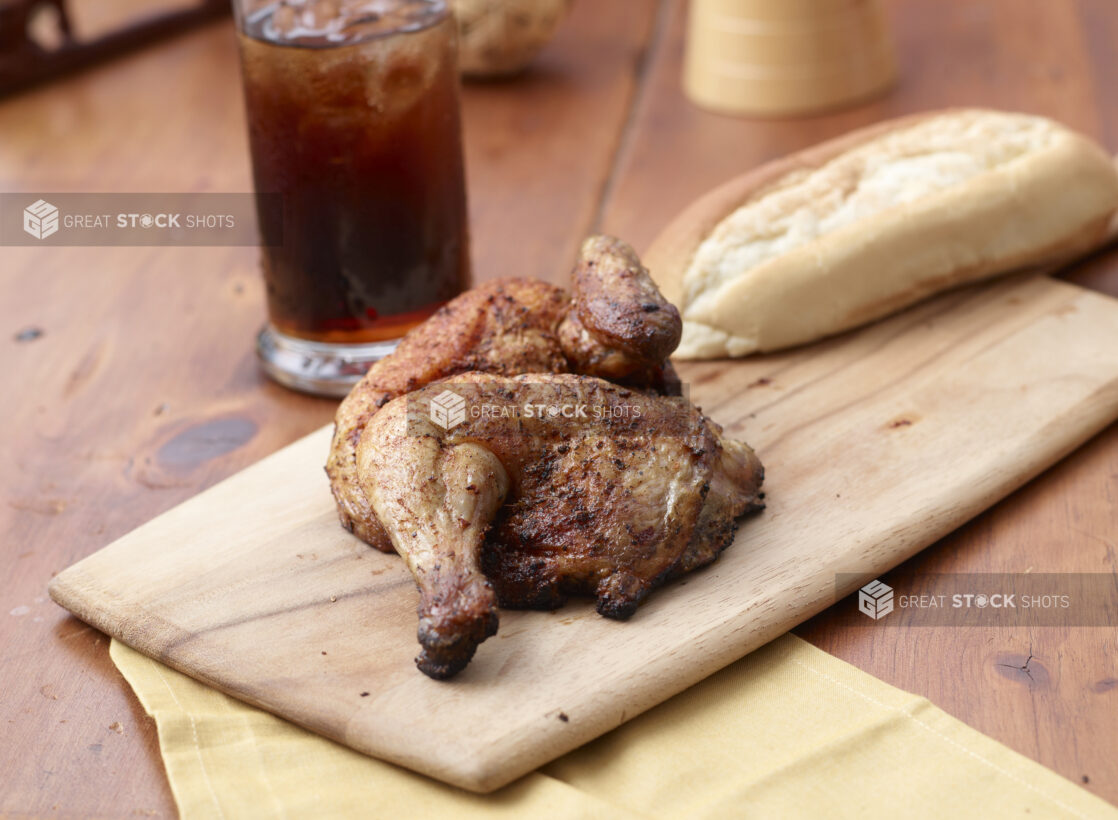 Rotisserie Chicken Leg on a Wooden Cutting Board with a Glass of Cola and Italian Bread on a Wooden Table in an Indoor Setting - Variation