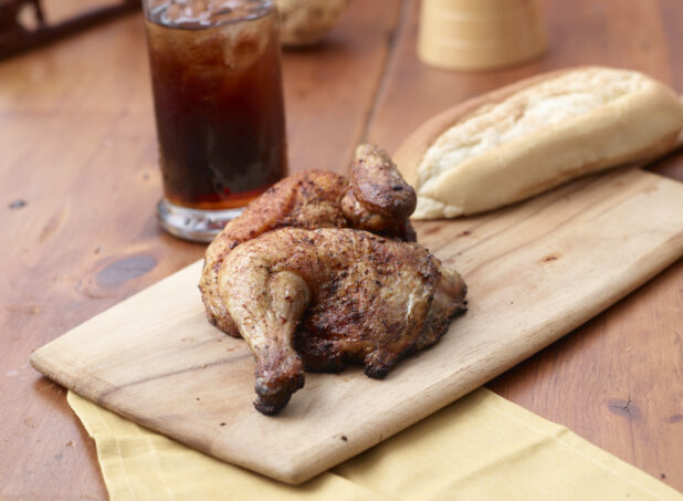Rotisserie Chicken Leg on a Wooden Cutting Board with a Glass of Cola and Italian Bread on a Wooden Table in an Indoor Setting - Variation