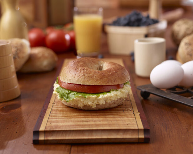 Egg Salad and Multigrain Bagel Sandwich with Lettuce and Tomato on a Wooden Platter Surrounded by Ingredients on Wooden Table