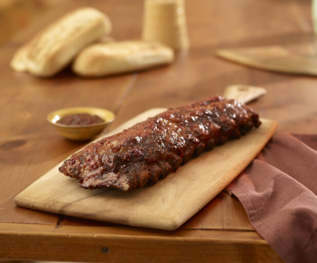 Whole Rack of Barbecue Ribs on a Wooden Cutting Board with a Side Dish of BBQ Sauce on a Wooden Table in an Indoor Setting