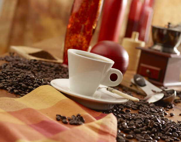 Hot Espresso Coffee in a White Ceramic Cup with Saucer and Spoon Surrounded by Whole Coffee Beans on a Wooden Table in an Indoor Setting