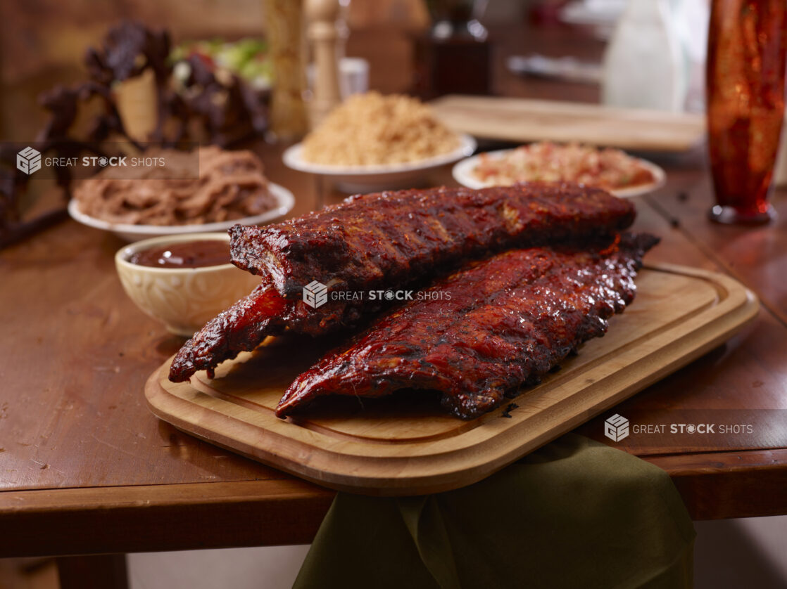 A Wooden Platter of Whole Racks of Ribs and Side Dishes of Spanish Rice, Tomato Salsa and Refried Beans on a Wooden Table in an Indoor Setting