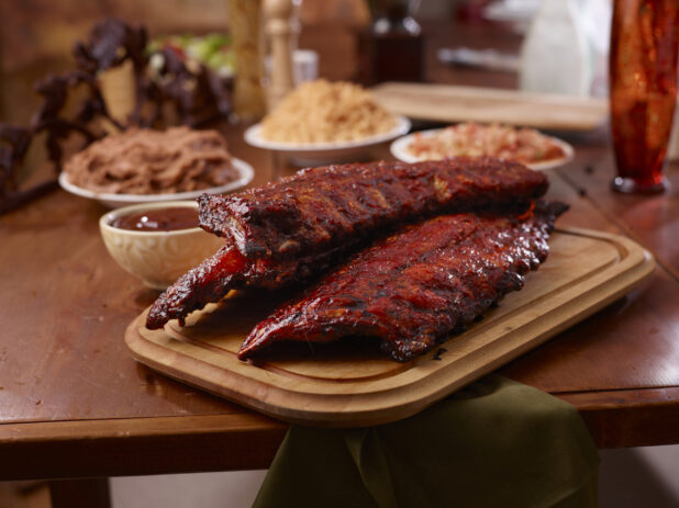 A Wooden Platter of Whole Racks of Ribs and Side Dishes of Spanish Rice, Tomato Salsa and Refried Beans on a Wooden Table in an Indoor Setting
