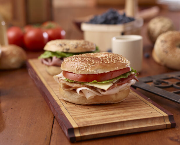 Oven-roasted Turkey, Bacon and Sesame Bagel Sandwich with Cheese, Lettuce and Tomato on a Wooden Platter with a Roast Beef Bagel Sandwich on a Wooden Table