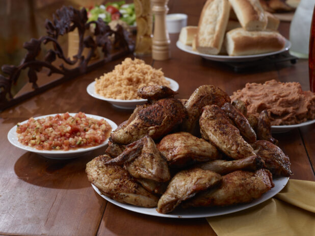 A Platter of Rotisserie Chicken Legs and Side Dishes of Spanish Rice, Tomato Salsa, Refried Beans and Baguettes on a Wooden Table in an Indoor Setting