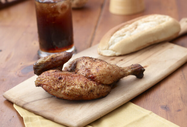 Rotisserie Chicken Leg on a Wooden Cutting Board with a Glass of Cola and Dinner Roll on a Wooden Table in an Indoor Setting