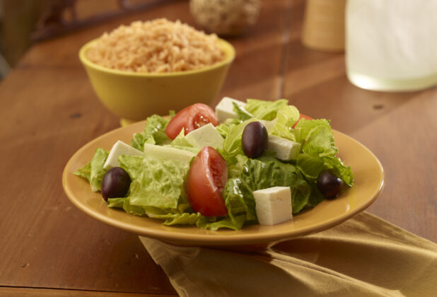 A Yellow Ceramic Bowl of Fresh Greek Salad with Kalamata Olives and Feta Cheese Cubes with a Bowl of Spanish Rice on a Wooden Table in an Indoor Setting