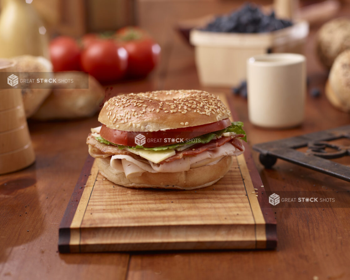 Oven-roasted Turkey, Bacon and Sesame Bagel Sandwich with Cheese, Lettuce and Tomato on a Wooden Platter Surrounded by Ingredients on Wooden Table