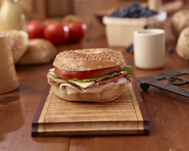 Oven-roasted Turkey, Bacon and Sesame Bagel Sandwich with Cheese, Lettuce and Tomato on a Wooden Platter Surrounded by Ingredients on Wooden Table