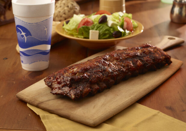 Whole Rack of Barbecue Ribs on a Wooden Cutting Board with a Take-Out Cup and a Bowl of Fresh Greek Salad on a Wooden Table in an Indoor Setting