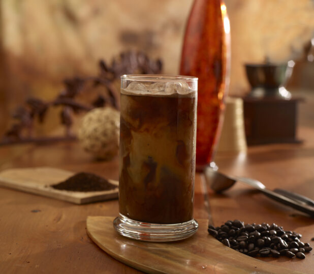 Iced Coffee and Cream in Glass Cup with Ice Cubes on a Wooden Table with Whole Coffee Beans and Ground Coffee in an Indoor Setting