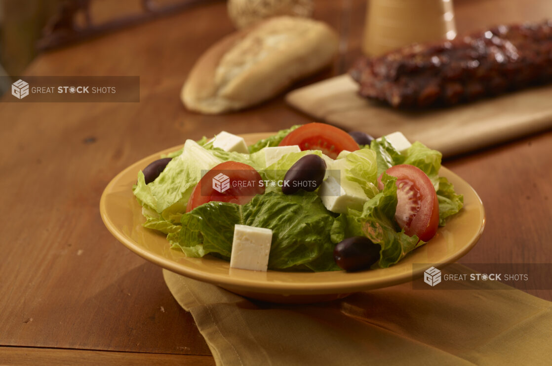 Side greek salad in the foreground with a rack of ribs and crusty bread in the background on a wooden table