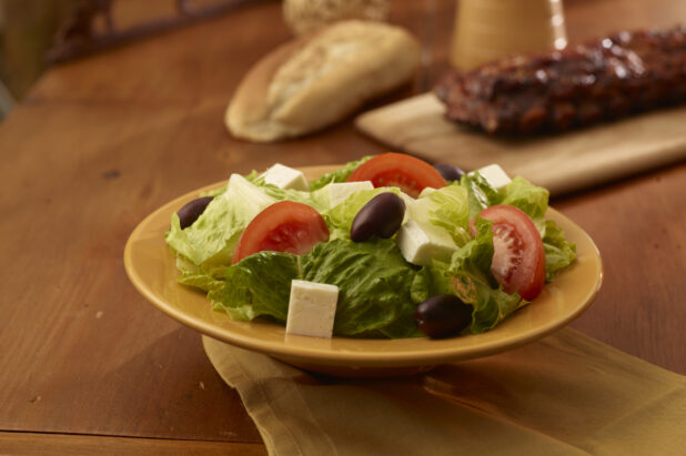 Side greek salad in the foreground with a rack of ribs and crusty bread in the background on a wooden table