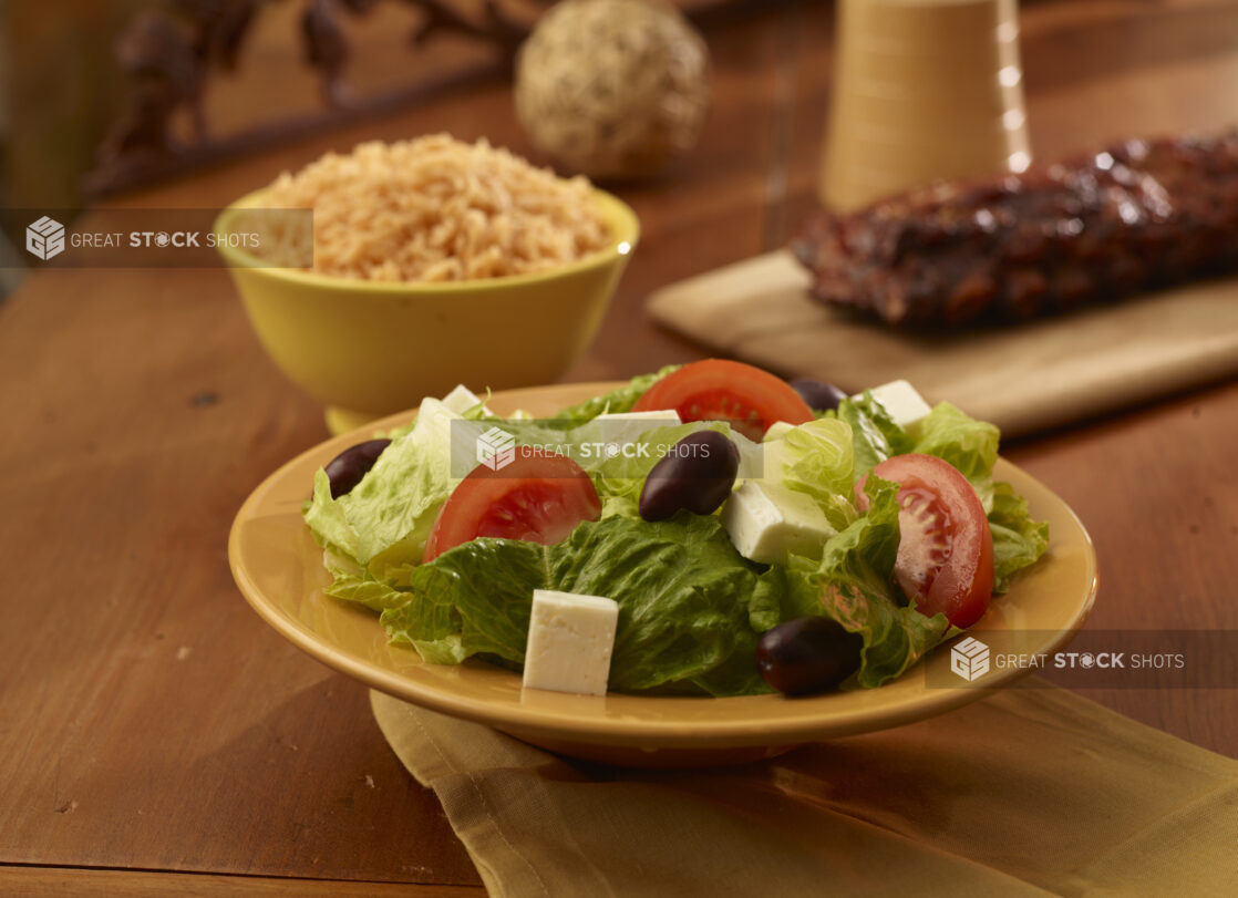 Side greek salad in the foreground with a rack of ribs, a side of rice and crusty bread in the background on a wooden table