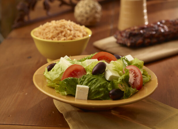 Side greek salad in the foreground with a rack of ribs, a side of rice and crusty bread in the background on a wooden table