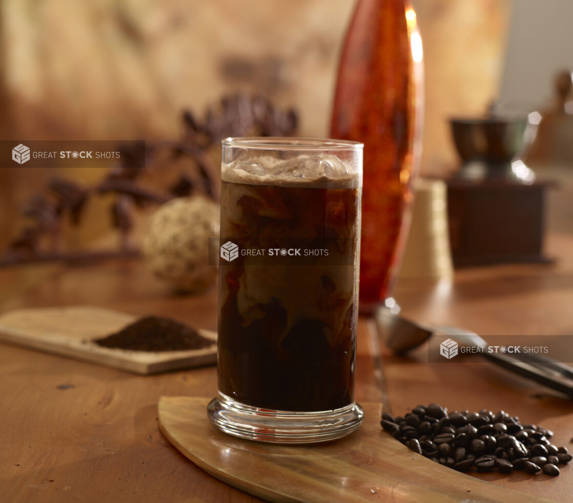 Iced coffee in a glass on a wooden board with coffee beans beside with ground coffee on a wooden board in the background