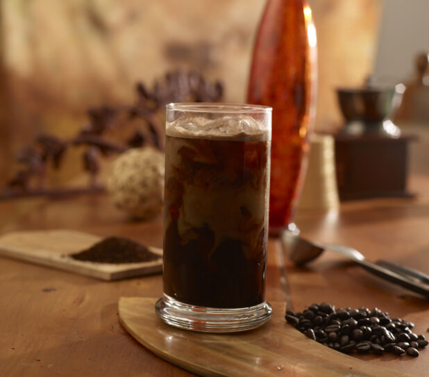Iced coffee in a glass on a wooden board with coffee beans beside with ground coffee on a wooden board in the background
