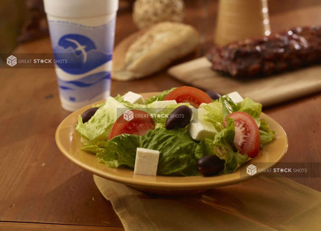 Side greek salad in the foreground with a rack of ribs, a beverage and crusty bread in the background on a wooden table