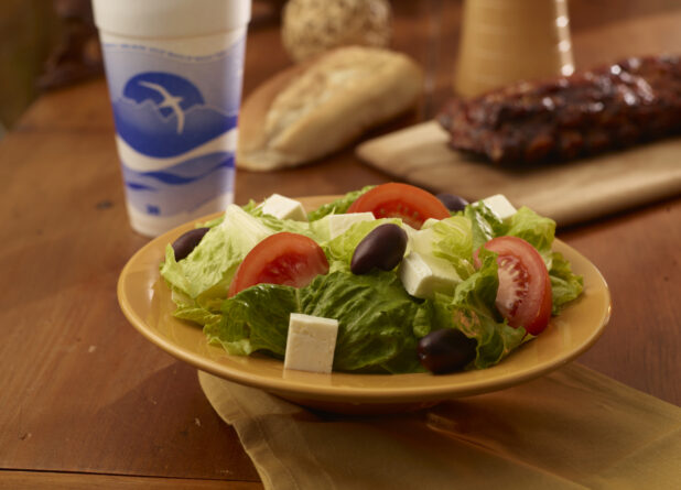 Side greek salad in the foreground with a rack of ribs, a beverage and crusty bread in the background on a wooden table