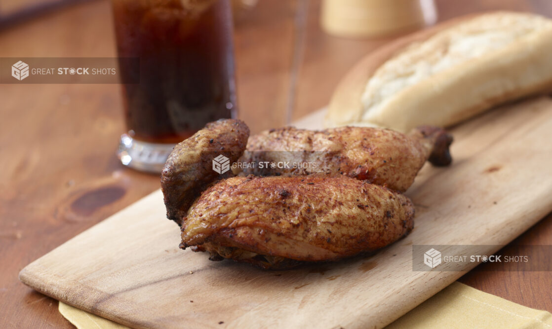 Roasted chicken on a wood board with crusty bread and a beverage in the background on a wooden background