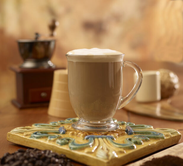 Cappuccino in a glass mug on a decorative trivet with coffee beans and ground coffee in the foreground with a coffee grinder in the background