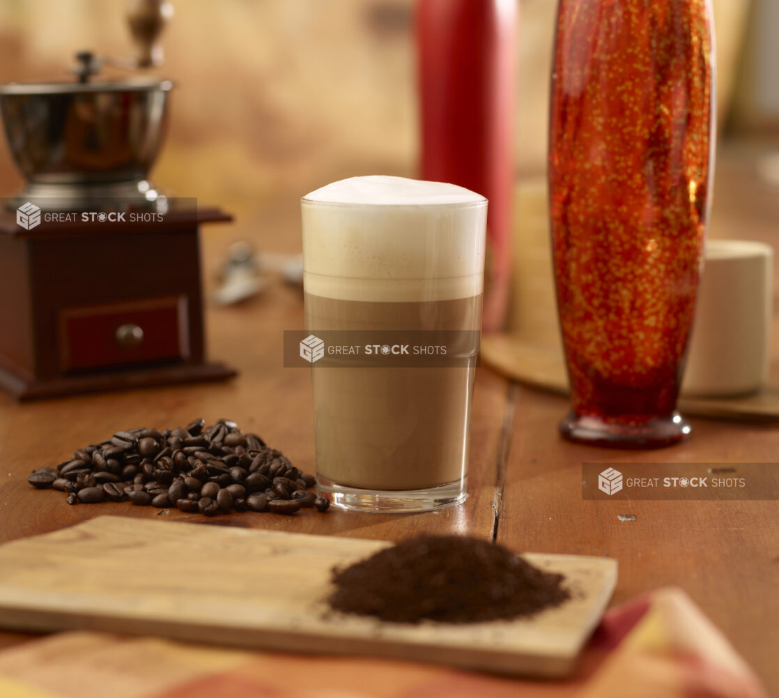 Café Latté with Milk Foam in a Tall Glass Cup with Whole Coffee Beans and Ground Coffee on a Wooden Table in an Indoor Setting