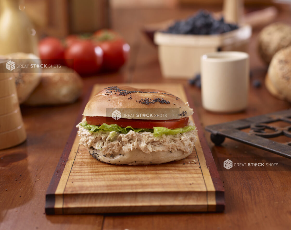 Tuna Salad and Poppy Seed Bagel Sandwich with Lettuce and Tomato on a Wooden Platter Surrounded by Ingredients on Wooden Table