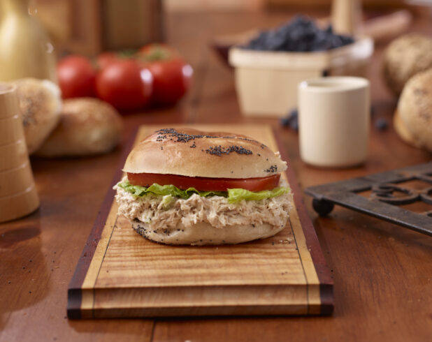 Tuna Salad and Poppy Seed Bagel Sandwich with Lettuce and Tomato on a Wooden Platter Surrounded by Ingredients on Wooden Table