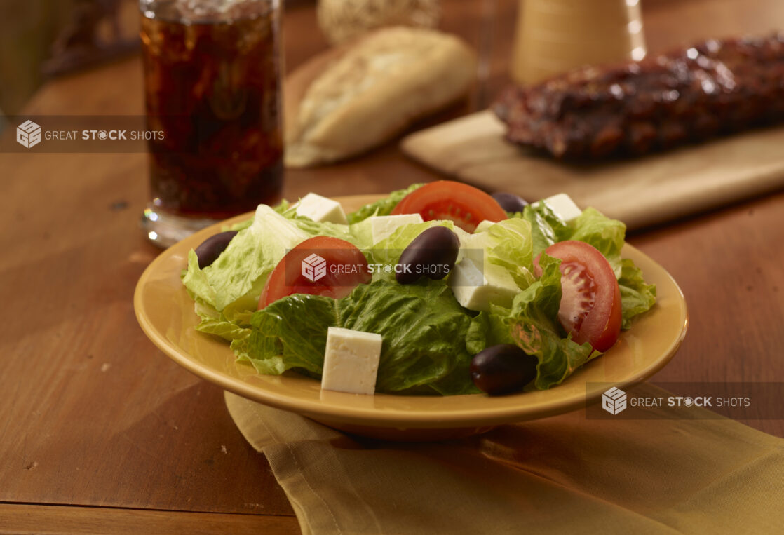 A Yellow Ceramic Bowl of Fresh Greek Salad with a Rack of Barbecue Ribs and a Glass of Cola on a Wooden Table in an Indoor Setting