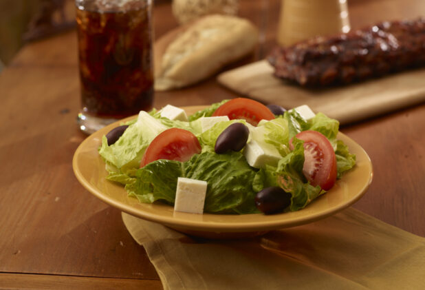 A Yellow Ceramic Bowl of Fresh Greek Salad with a Rack of Barbecue Ribs and a Glass of Cola on a Wooden Table in an Indoor Setting