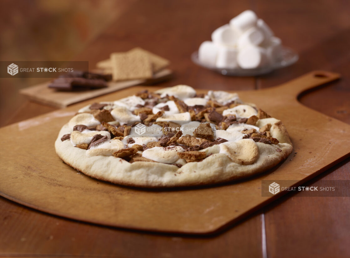 Whole Baked S'Mores Pizza on a Wooden Pizza Peel with Chocolate, Graham Crackers and Marshmallows in the Background on a Wooden Table