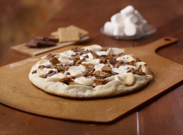 Whole Baked S'Mores Pizza on a Wooden Pizza Peel with Chocolate, Graham Crackers and Marshmallows in the Background on a Wooden Table