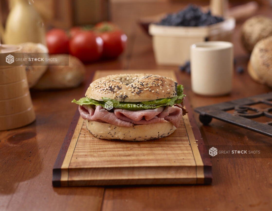 Sliced Delicatessen Roast Beef and Everything Bagel Sandwich with Lettuce and Tomato on a Wooden Platter Surrounded by Ingredients on Wooden Table