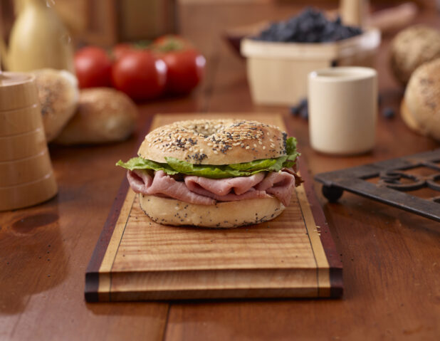 Sliced Delicatessen Roast Beef and Everything Bagel Sandwich with Lettuce and Tomato on a Wooden Platter Surrounded by Ingredients on Wooden Table