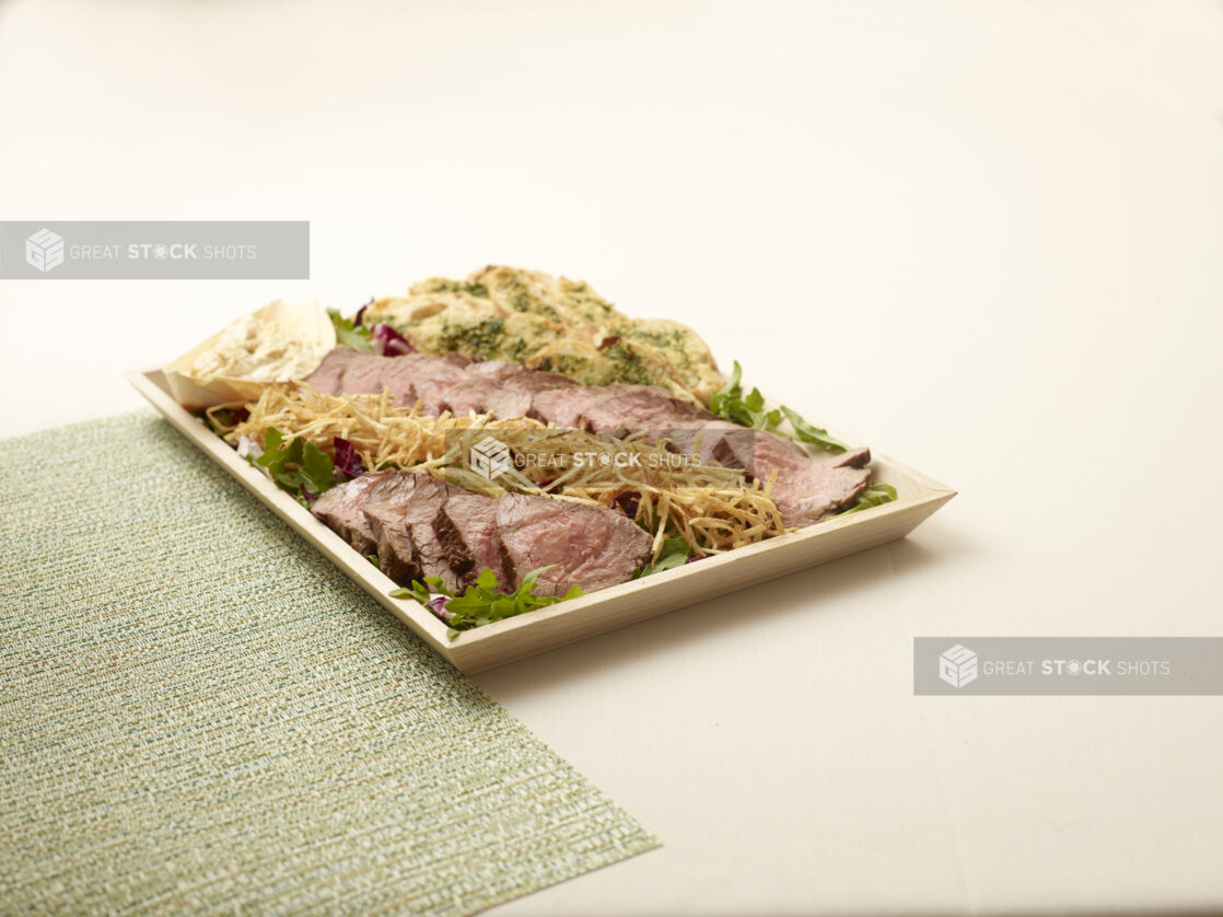 Large Wood Tray with Sliced Grilled Pork Tenderloin on a Bed of Spring Mix Salad with Garlic Bread Crostini and Fried Onions on a Beige Surface with a Green Placemat - for Catering